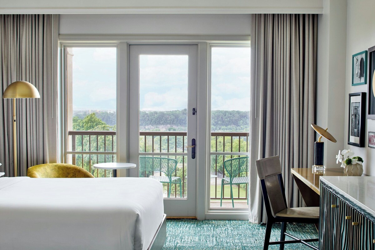 A hotel room with a bed, a desk, a chair, a lamp, and a view of greenery through large windows.