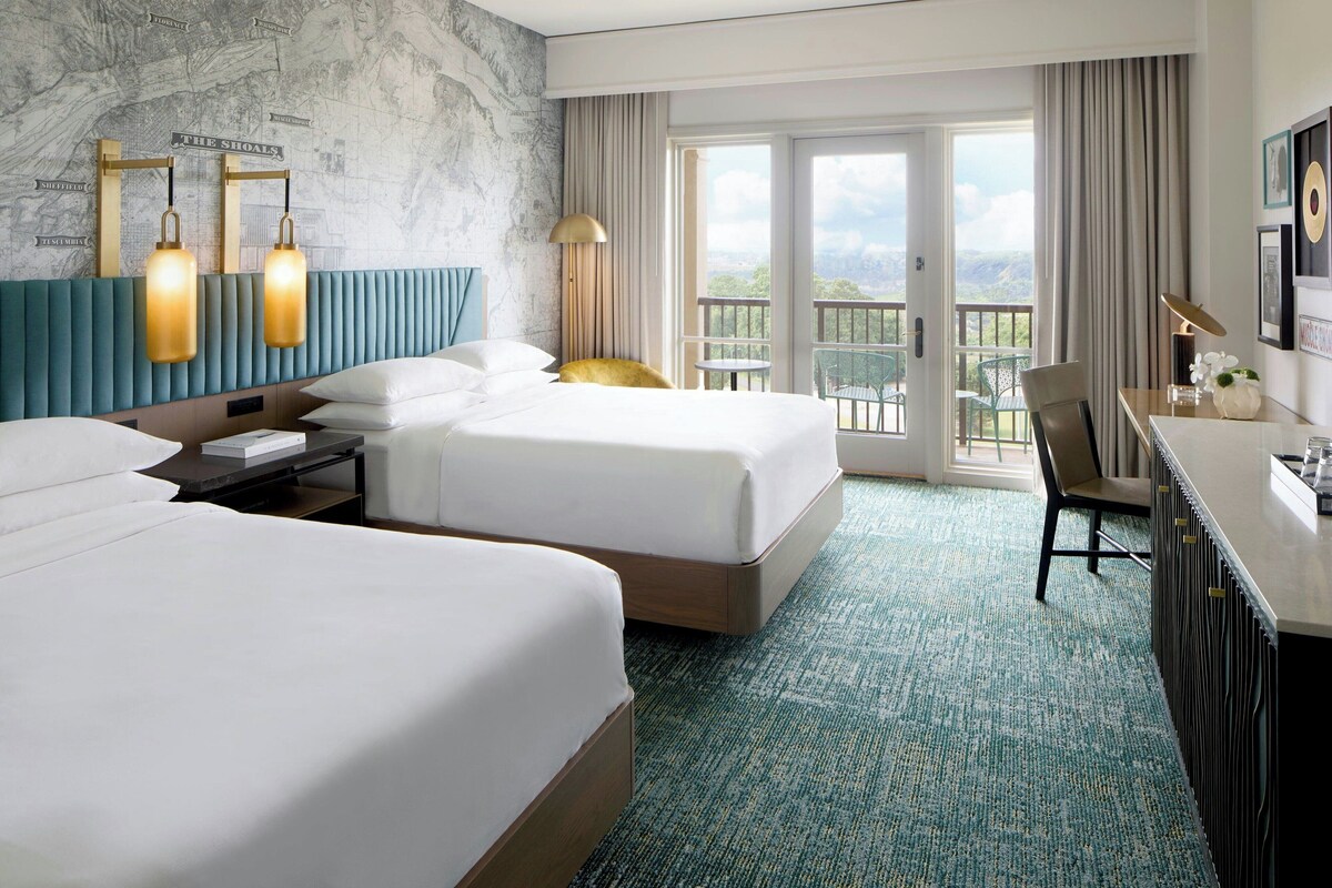 A hotel room with two beds, a desk, a chair, a balcony with a view, and a wall with a map.
