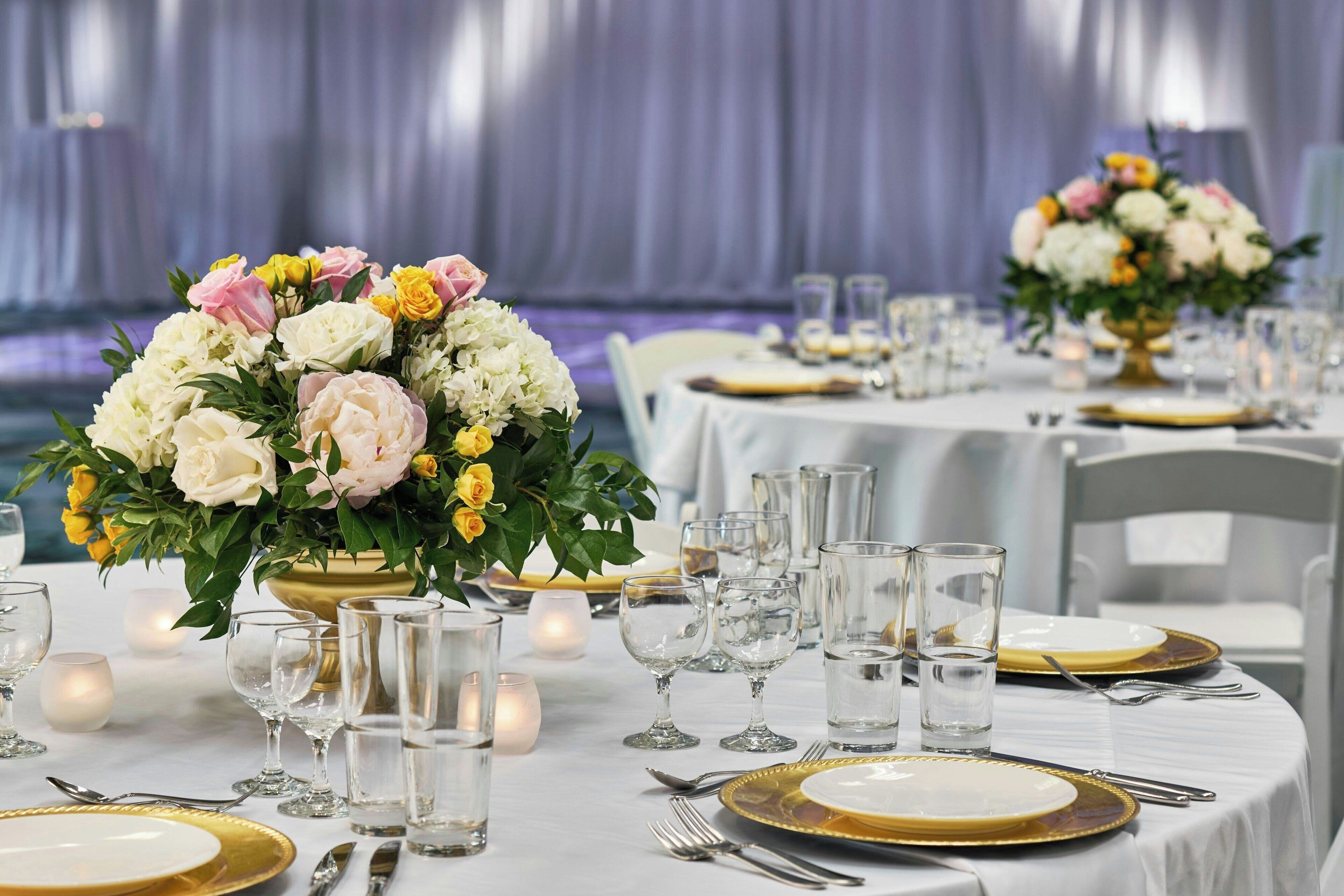 A banquet hall with round tables set for dining, each with gold-rimmed plates and clear glassware.