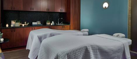 A spa room with two beds, wooden cabinets, a sink, and a small table with plants and candles.