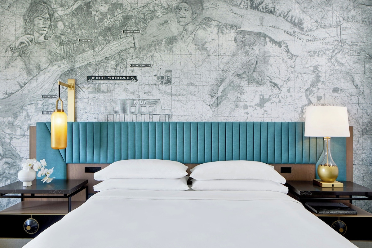 Habitación de hotel con una cama grande, cabecero tapizado en color verde azulado, pared con motivos de mapa y dos mesitas de noche con lámparas.