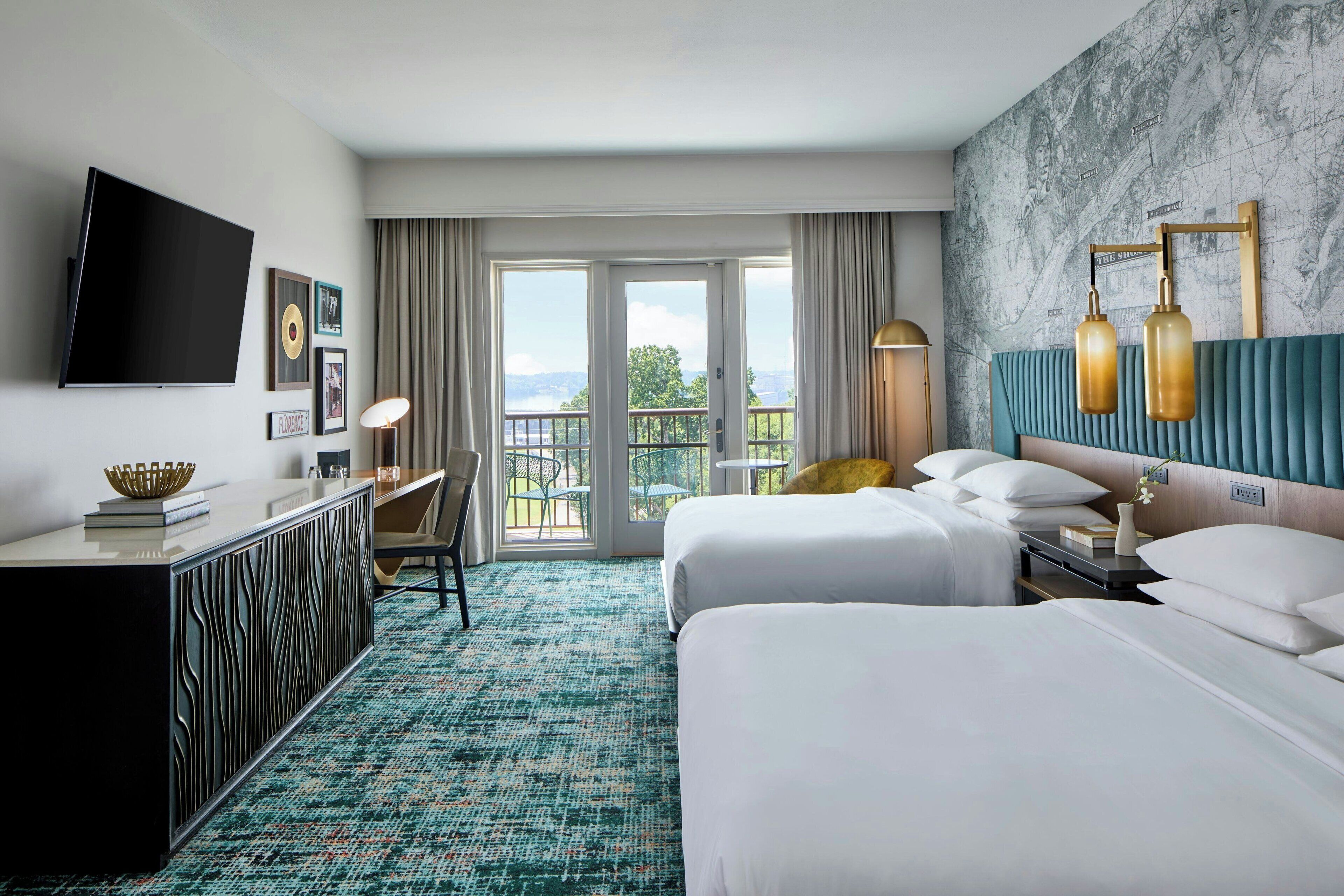A hotel room with two beds, a desk, a TV, and a balcony with a view.