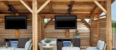 Een overdekt terras met rieten meubels, twee televisies bevestigd aan houten balken en een plafondventilator.