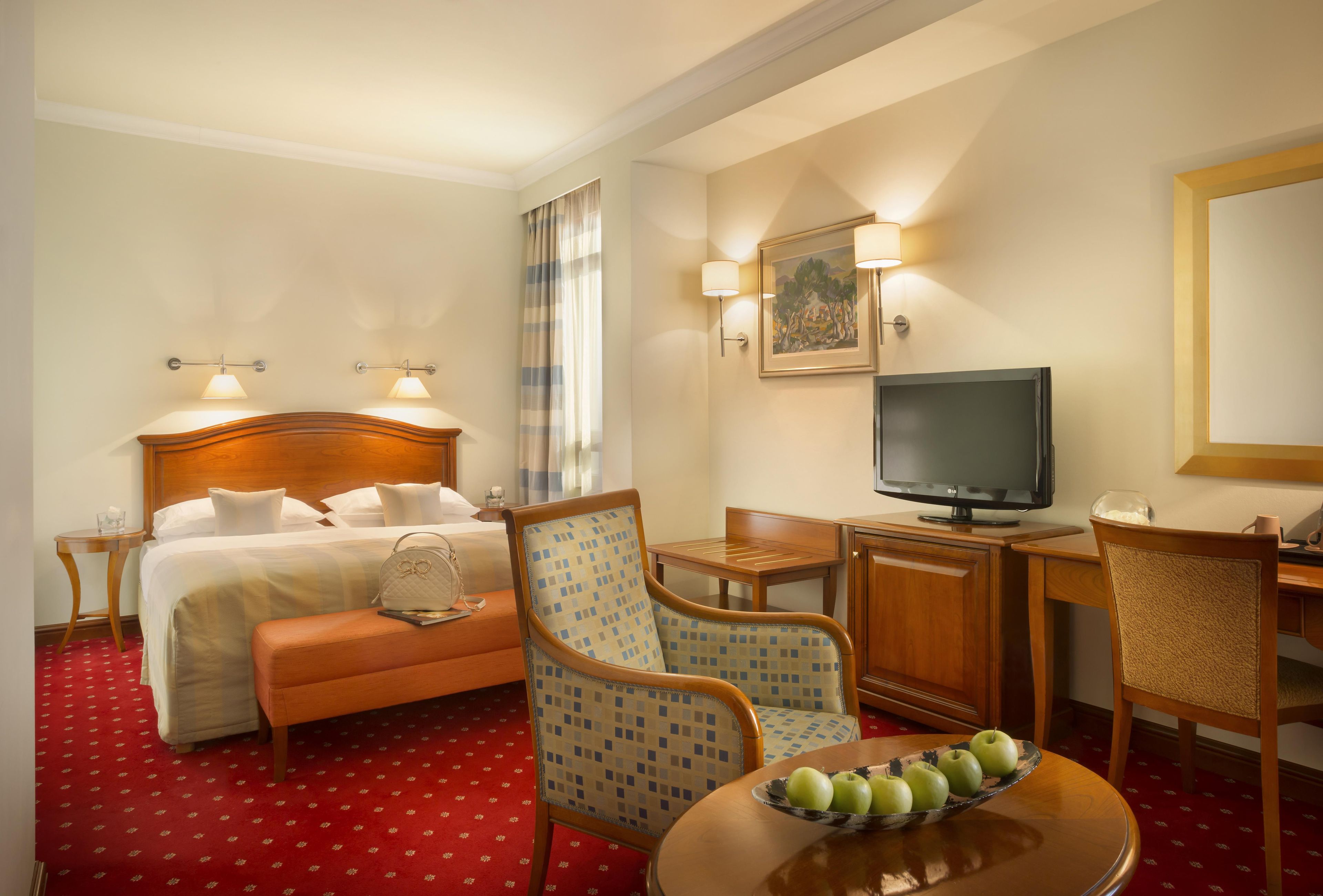 A hotel room with a bed, a desk with a television, a chair, a table with apples, and a painting on the wall.