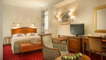 A hotel room with a bed, a desk with a television, a chair, a table with apples, and a painting on the wall.