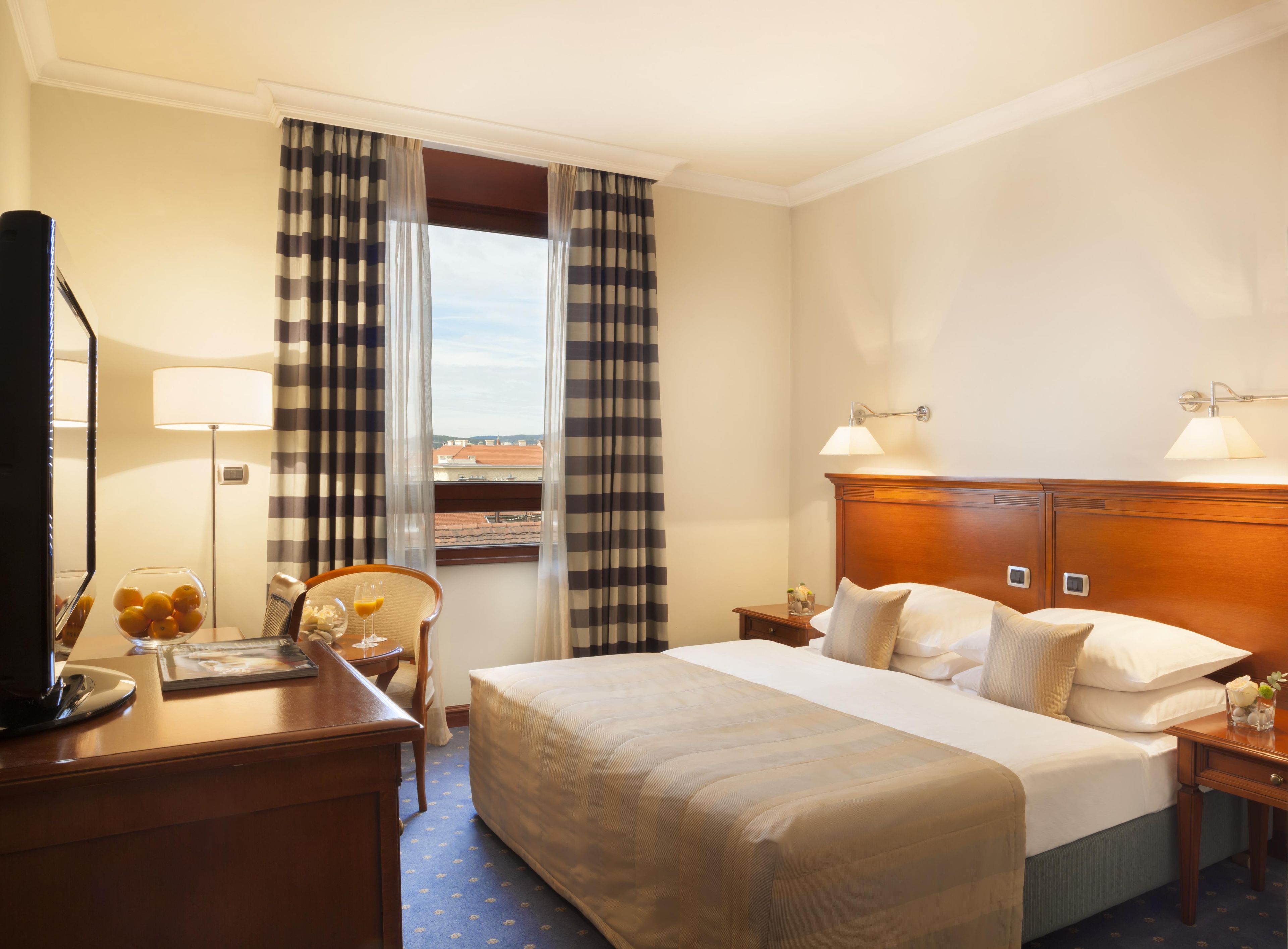 A hotel room with a bed, bedside lamps, a desk with a lamp, a chair, a TV, and a window with curtains.