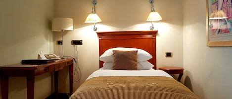 A hotel room with a bed, two bedside lamps, a desk with a telephone, and a painting on the wall.