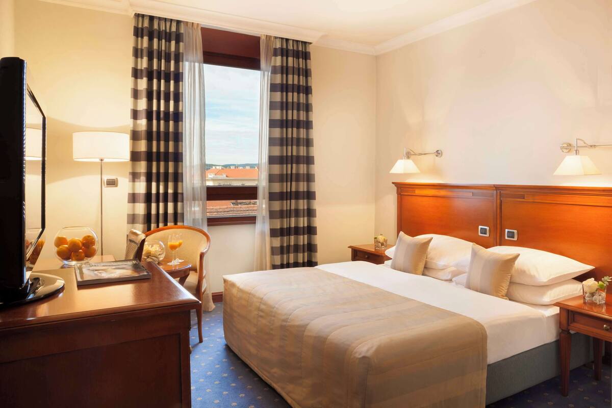 A hotel room with a bed, bedside lamps, a desk with a lamp, a chair, a bowl of fruit, and a window with curtains.