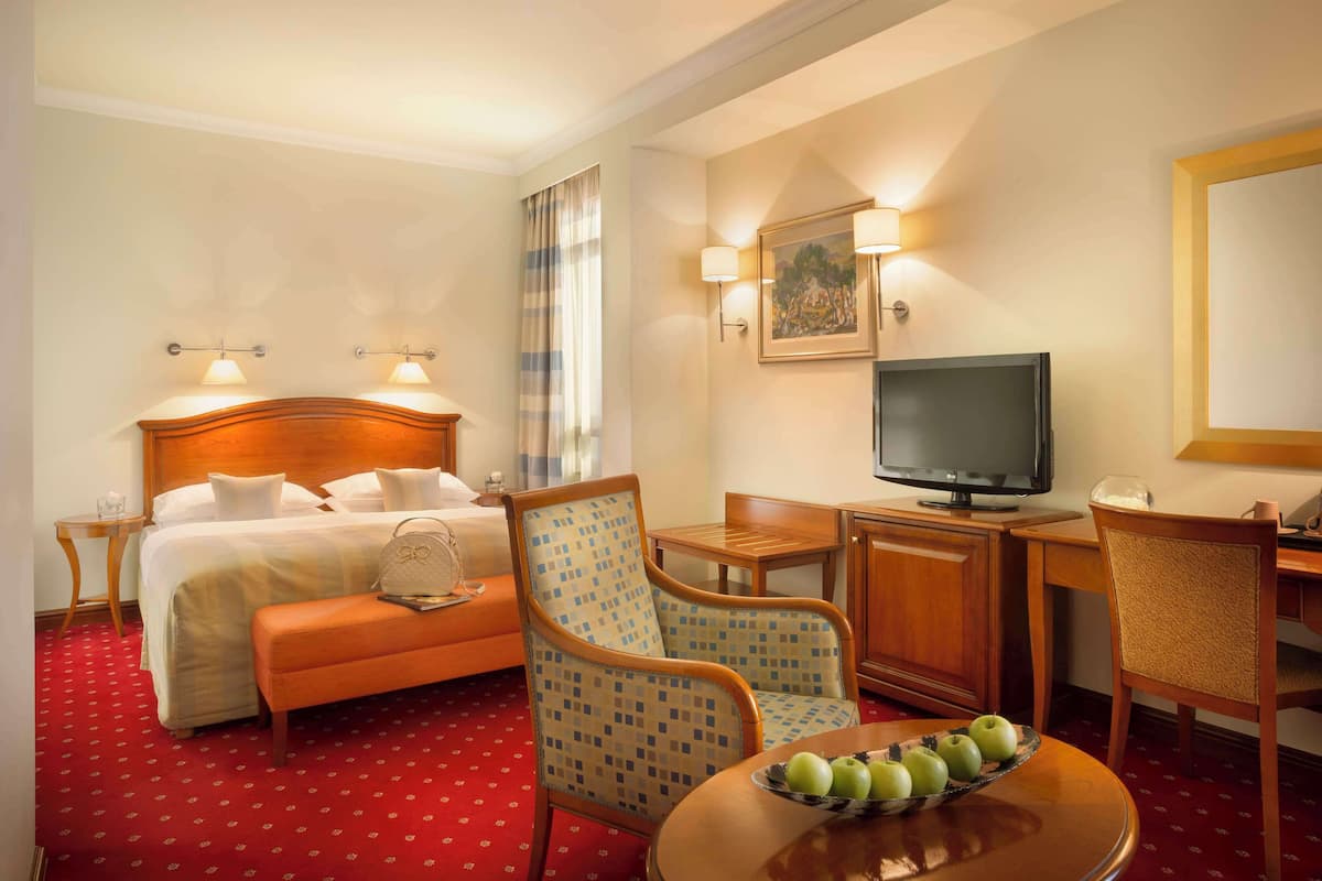 A hotel room with a bed, a desk with a television, a chair, a table with apples, and a painting on the wall.