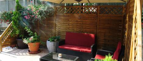 Un patio al aire libre con cerca de madera, un sofá rojo, una mesa de centro de vidrio y plantas en macetas.