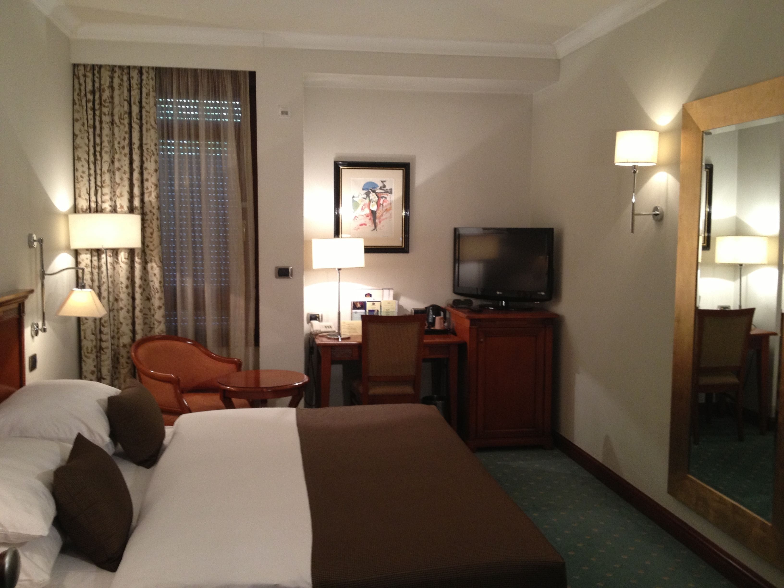 A hotel room with a bed, a desk with a television, a chair, and a large mirror.