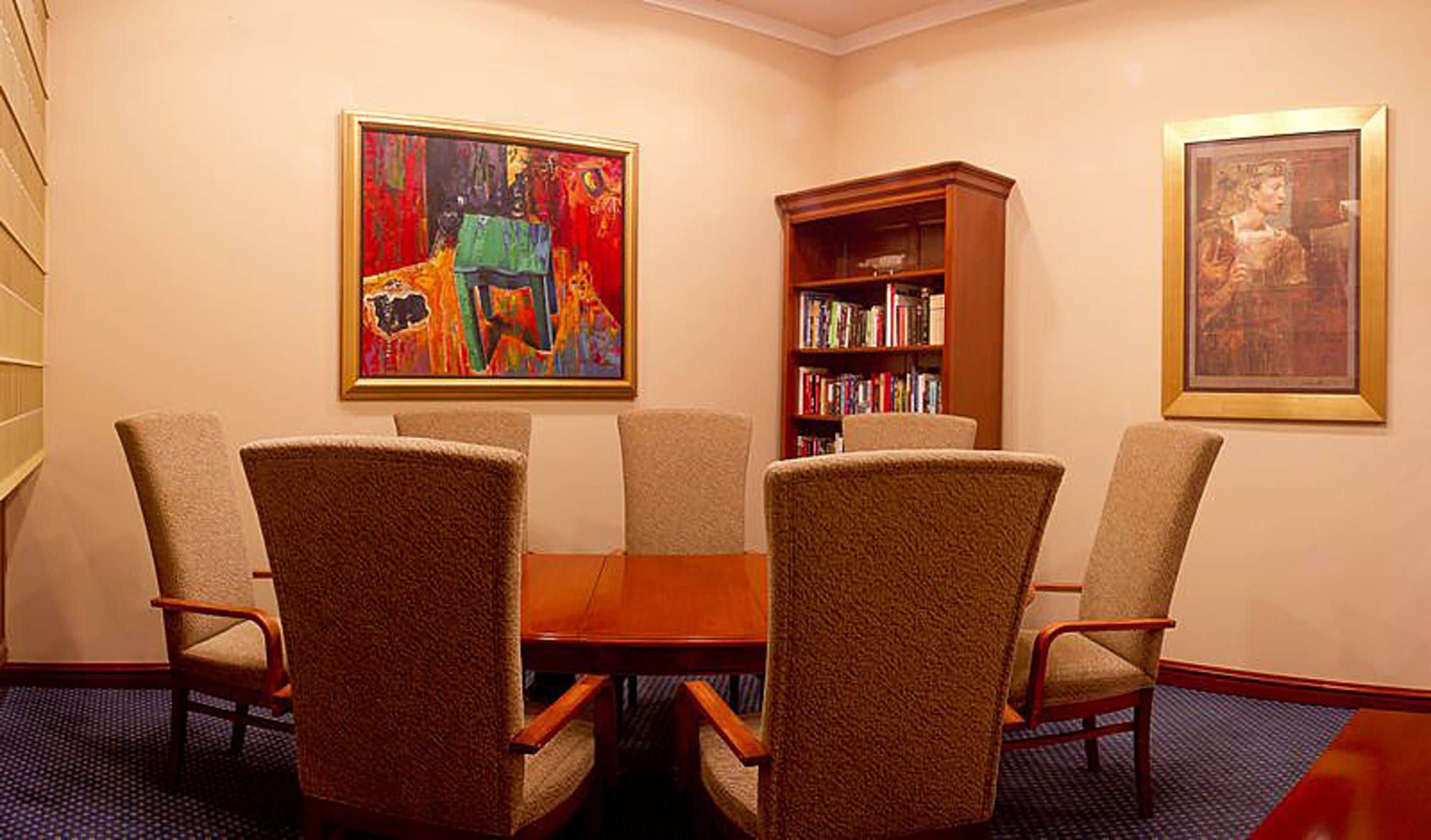 A conference room with a round table, six chairs, a bookshelf, and two framed artworks on the walls.