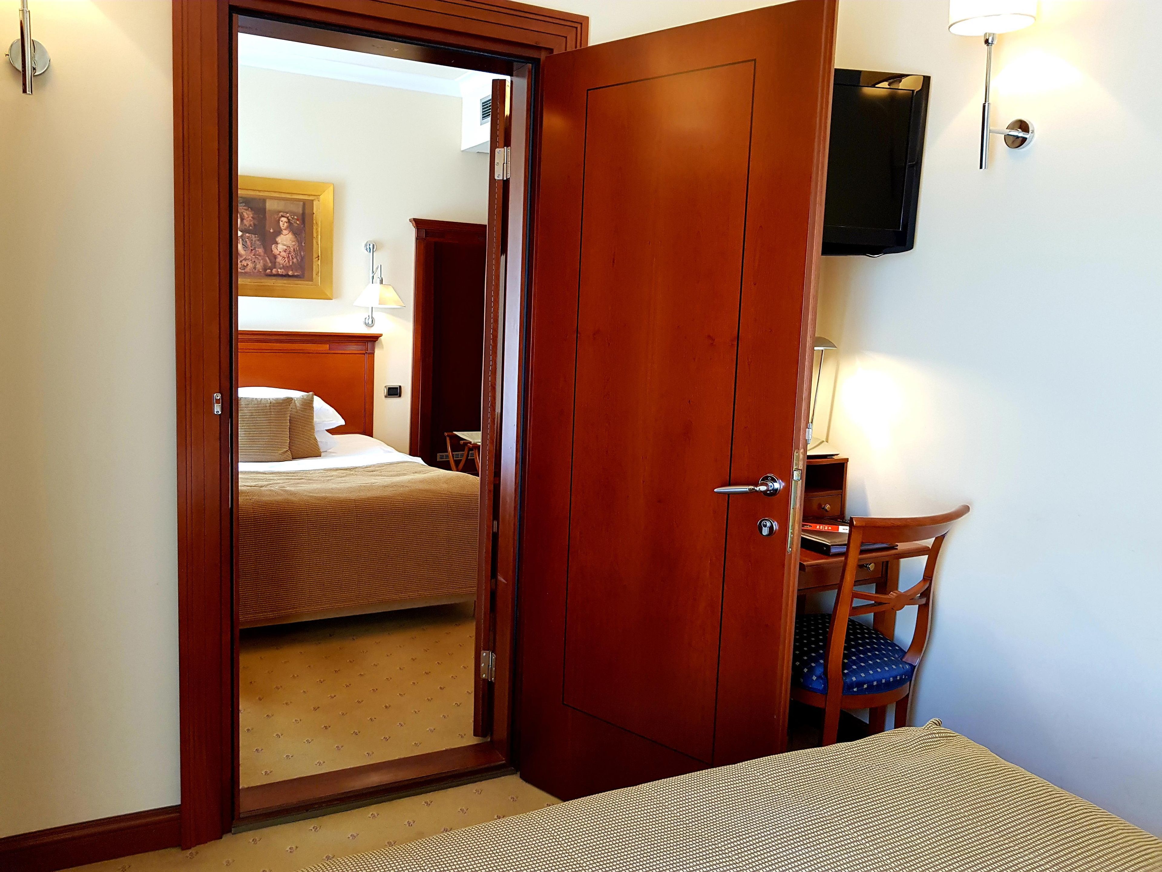 Habitación de hotel con puerta de madera, una cama, un escritorio con silla y un televisor montado en la pared.