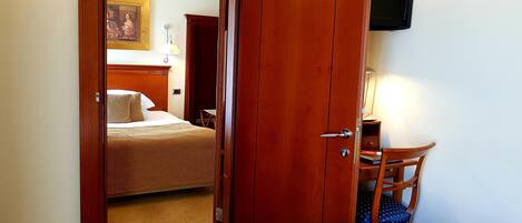Habitación de hotel con puerta de madera, una cama, un escritorio con silla y un televisor montado en la pared.