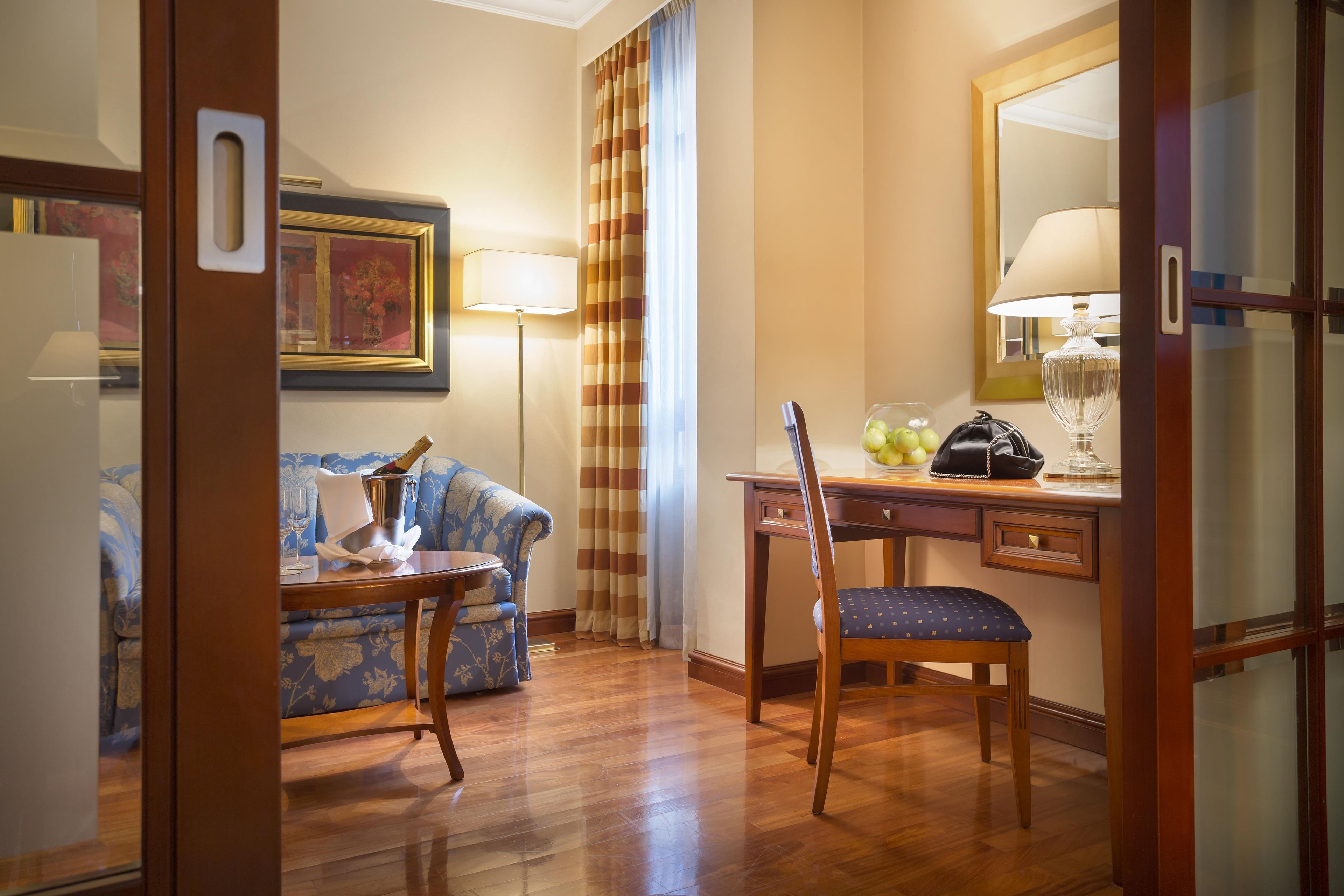 A hotel room with a wooden floor, a sofa, a desk with a chair, a lamp, and a framed picture on the wall.