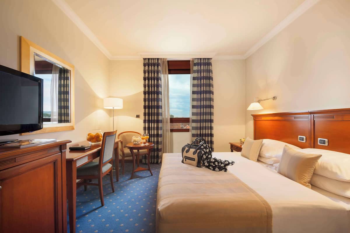 A hotel room with a bed, a desk with a television, a chair, a table with a glass of orange juice, and curtains.