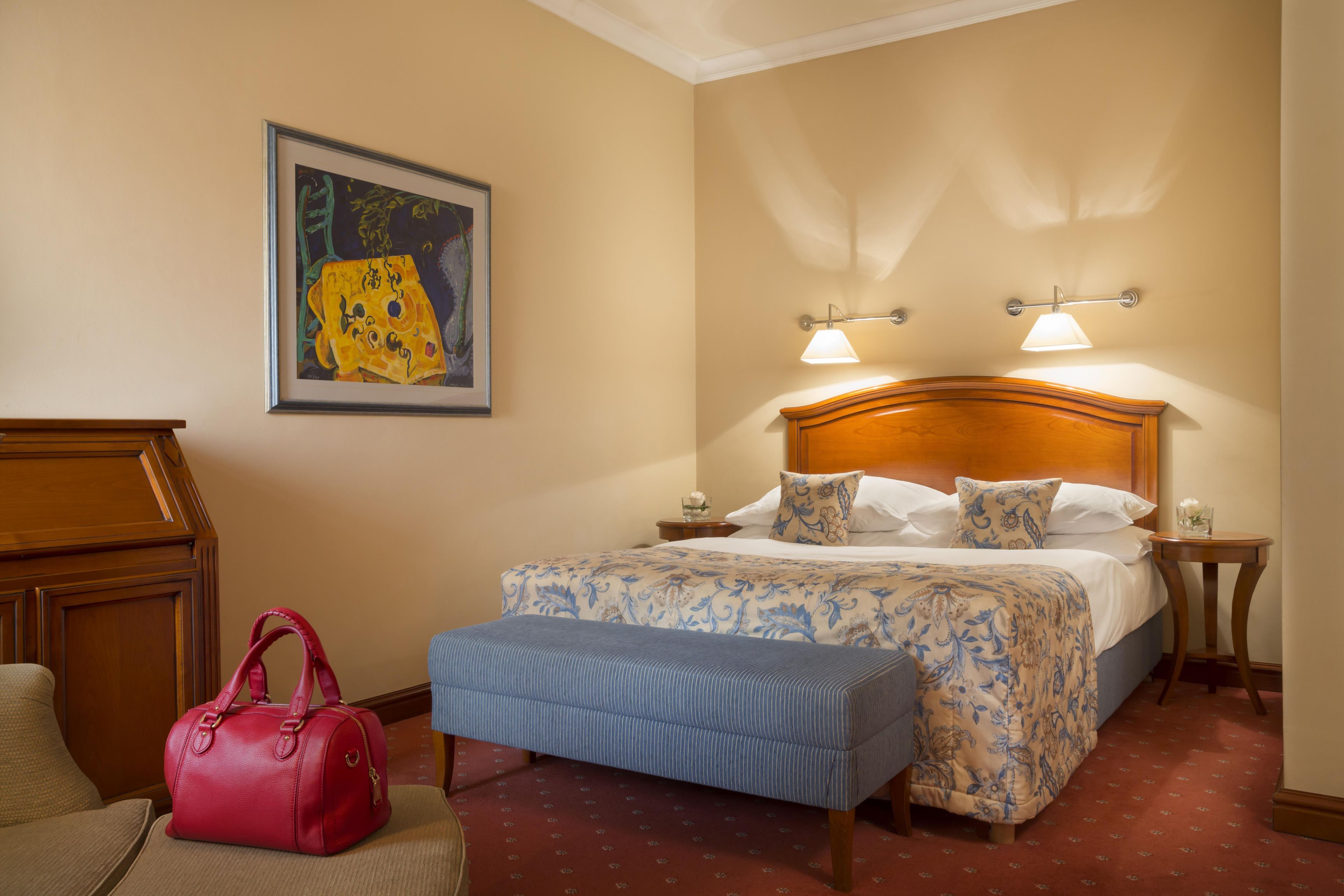 Une chambre d’hôtel avec un lit, une table de chevet, une lampe, un sac à main rouge et un tableau accroché au mur.