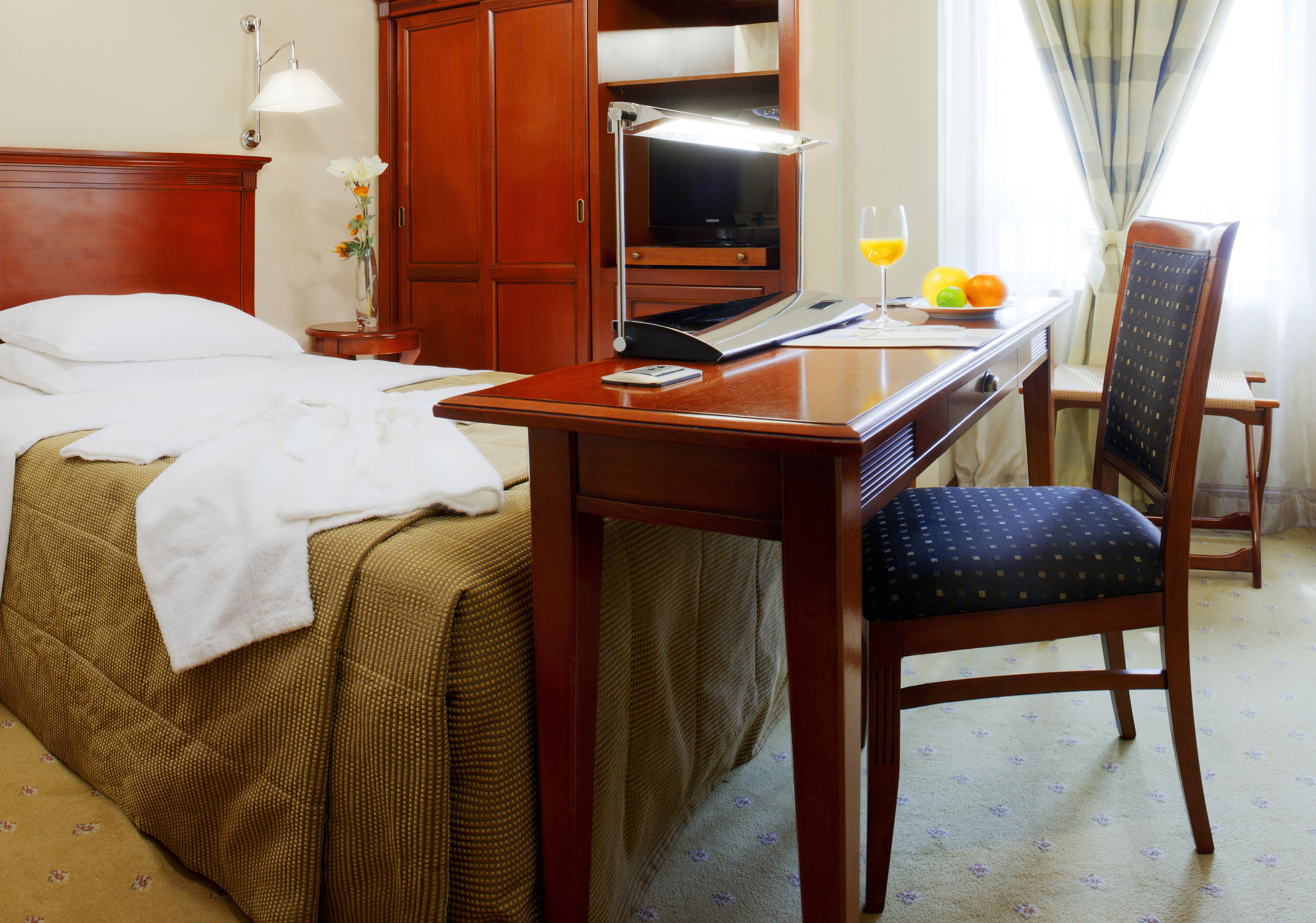 Habitación de hotel con cama, escritorio con lámpara, silla y una pequeña mesa con frutas y un vaso de jugo de naranja.