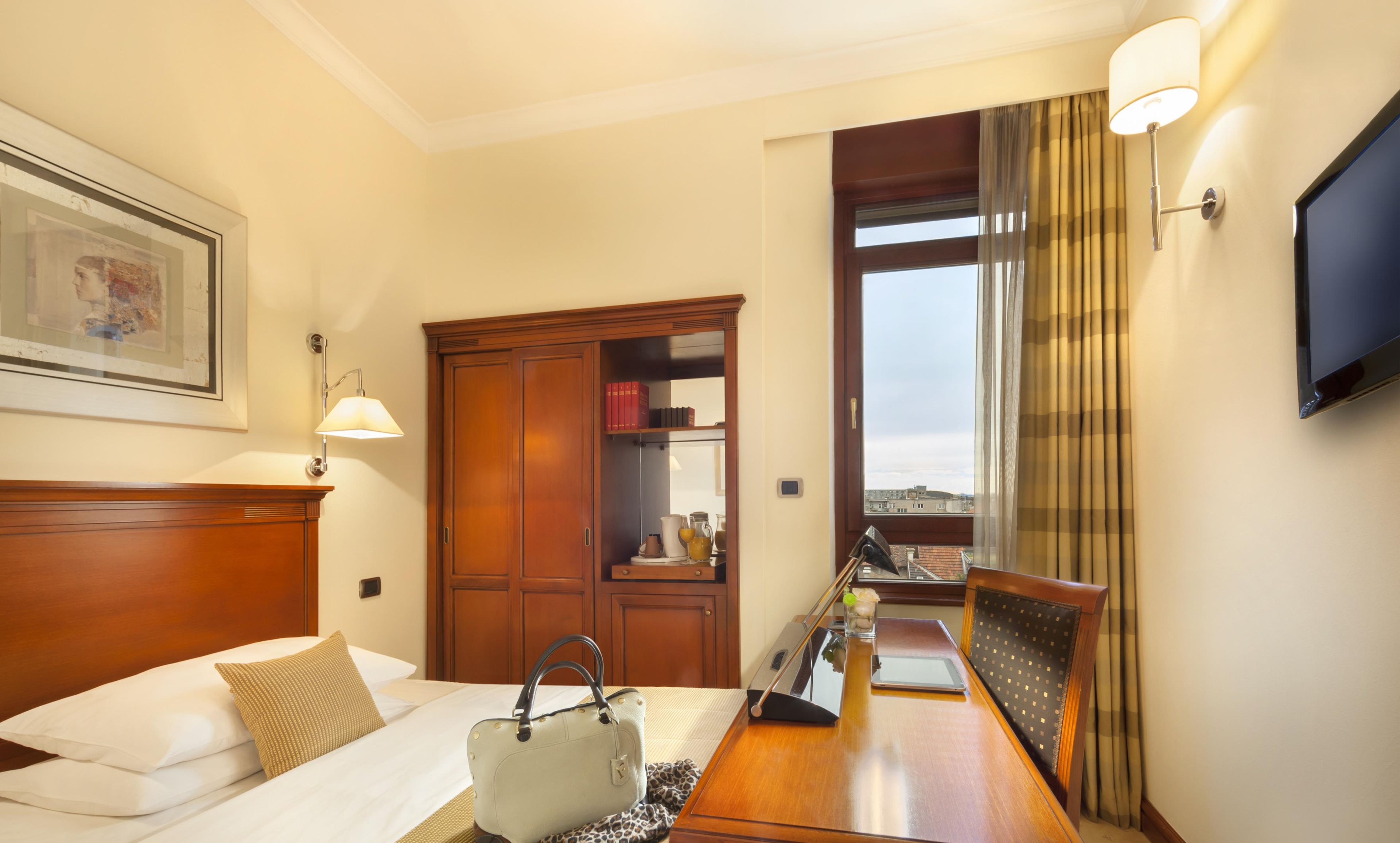 A hotel room with a bed, bedside table, lamp, and a view of the city through a window.
