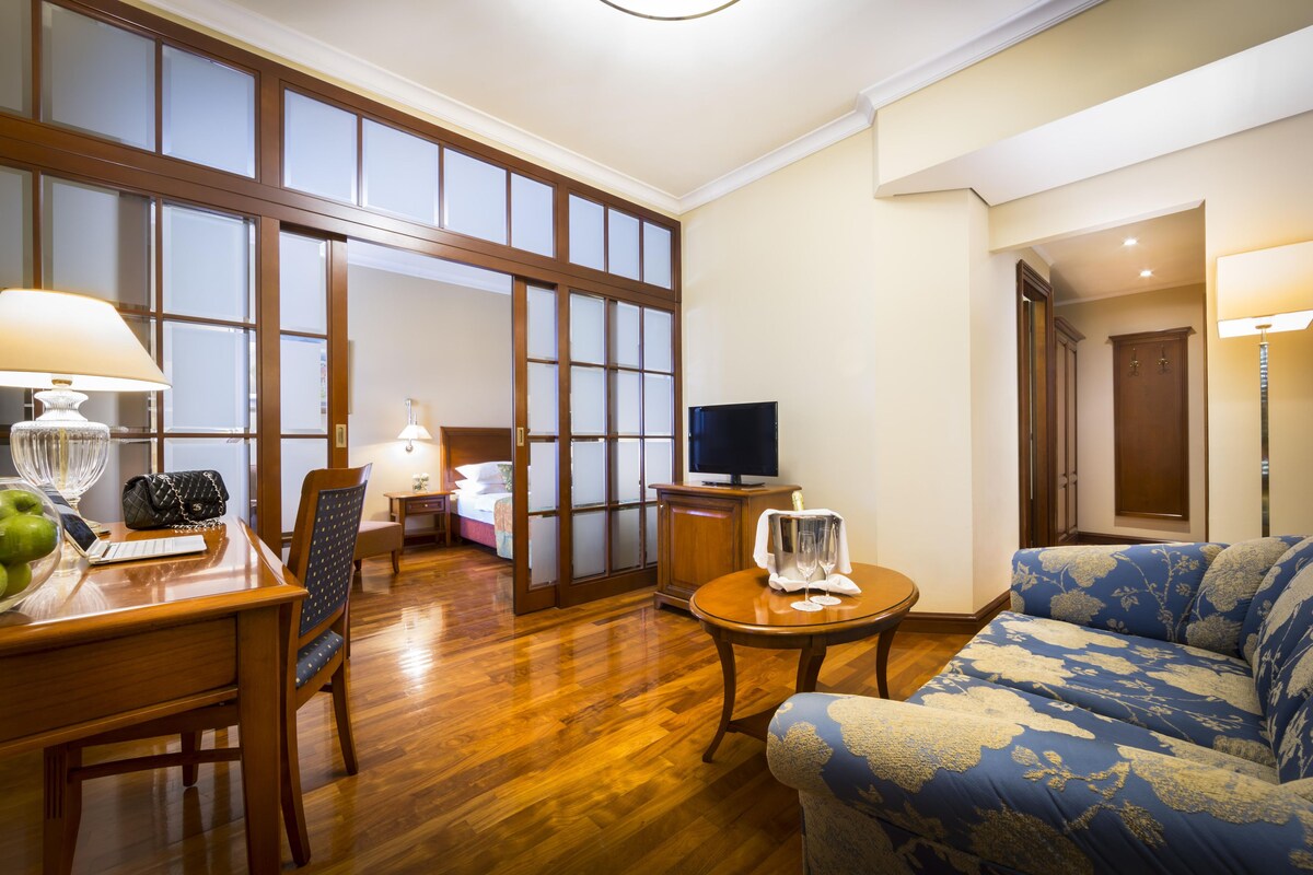 Habitación de hotel con piso de madera, un sofá, una mesa de comedor con sillas, un televisor y un ventanal con paneles de vidrio esmerilado.