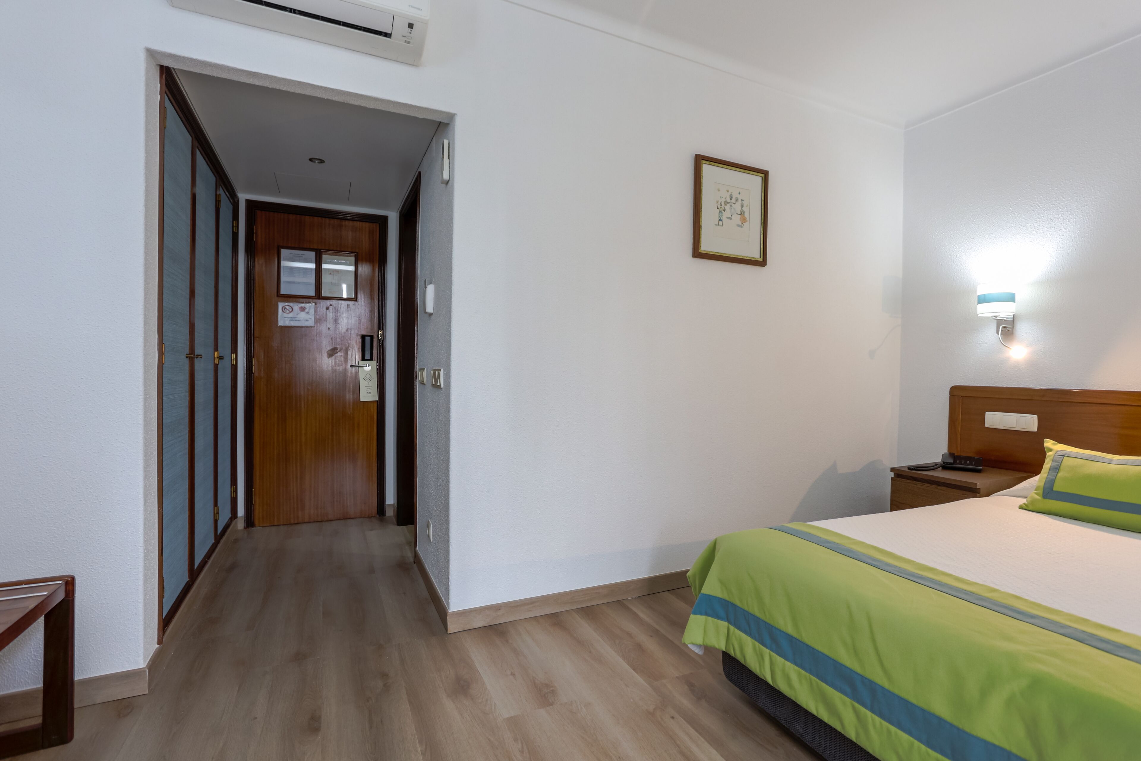 A hotel room with a wooden door, a bed with a green and blue striped bedspread, a bedside table with a lamp, and a framed picture on the wall.