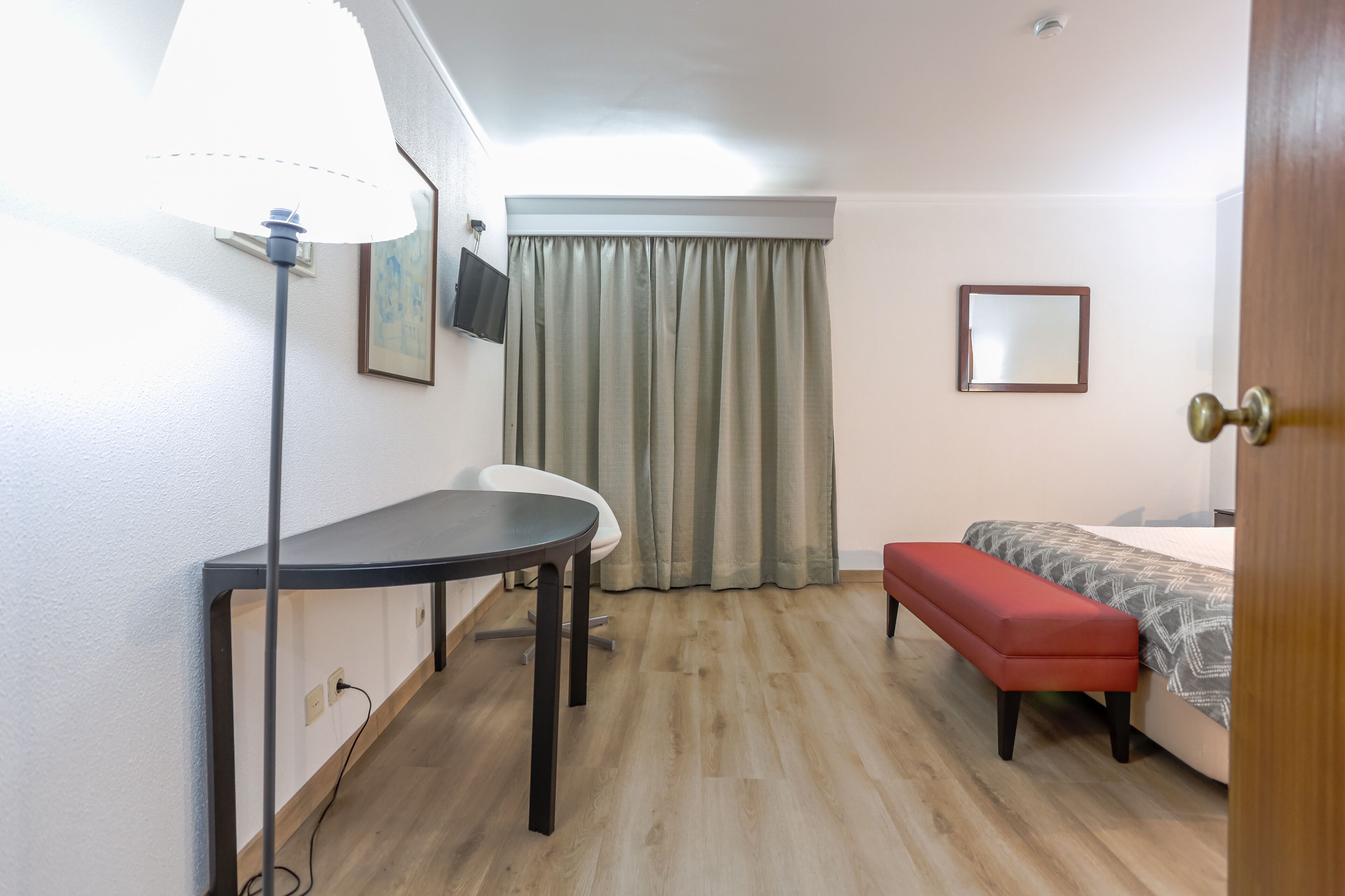 Habitación de hotel con suelo de madera, un escritorio con lámpara, una silla, una cama con un banco rojo y una ventana con cortinas.