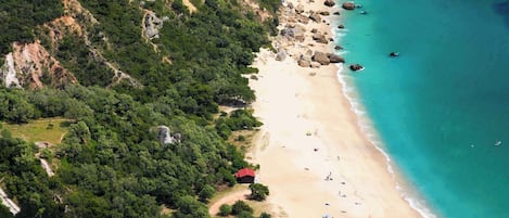 An aerial view of a coastal area with a sandy beach, dense greenery, and a clear blue sea.
