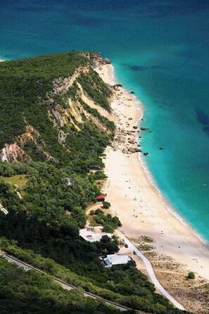 An aerial view of a coastal area with a sandy beach, dense greenery, and a clear blue sea. - Hotel Aranguês (Setubal)