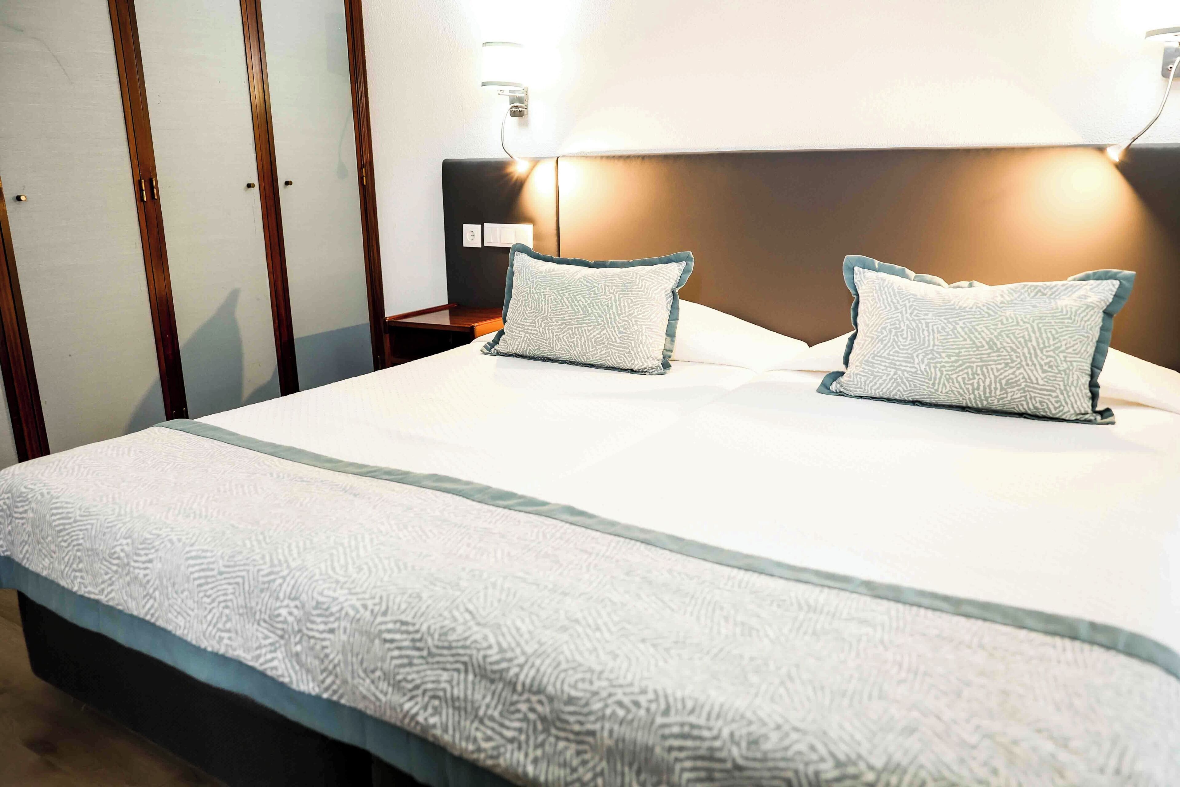 A hotel room with a large bed, two pillows, a nightstand, and a wall-mounted lamp.