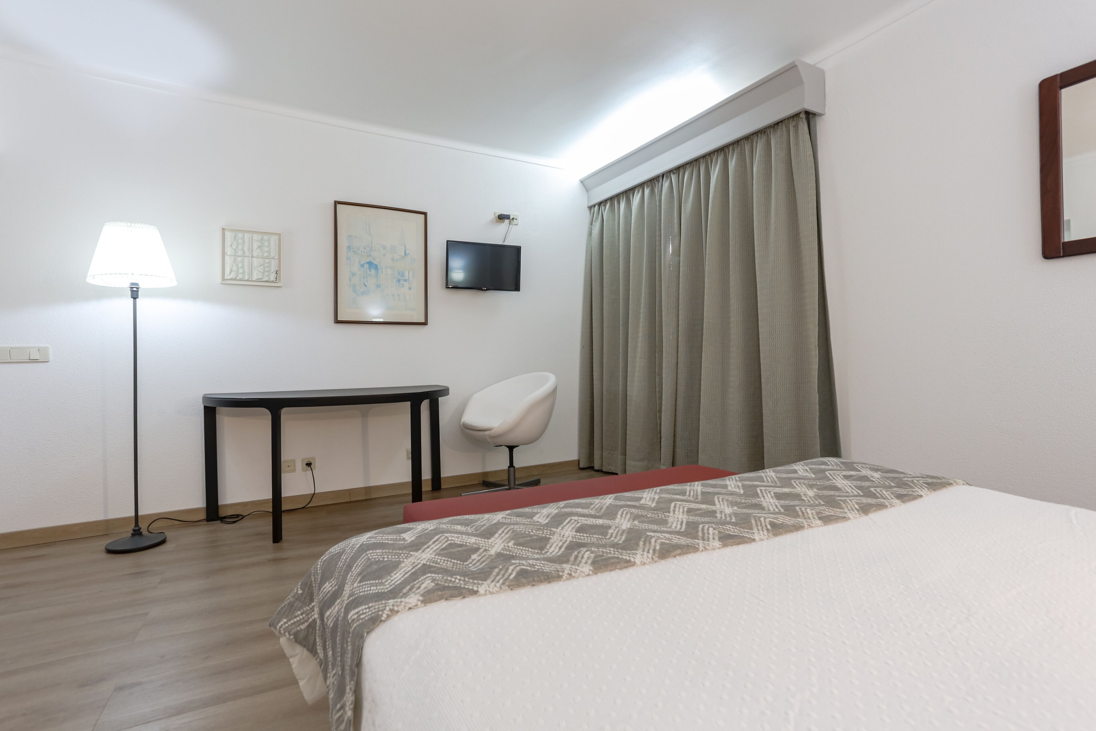 A hotel room with a bed, a desk, a chair, a TV, and a lamp.