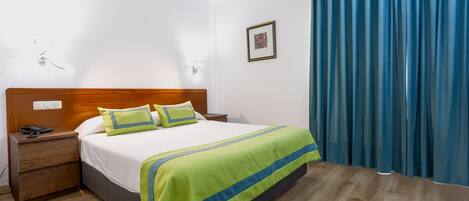 Habitación de hotel con cama de cabecera de madera, colcha a rayas verde y azul, mesita de noche con teléfono y cortina azul.