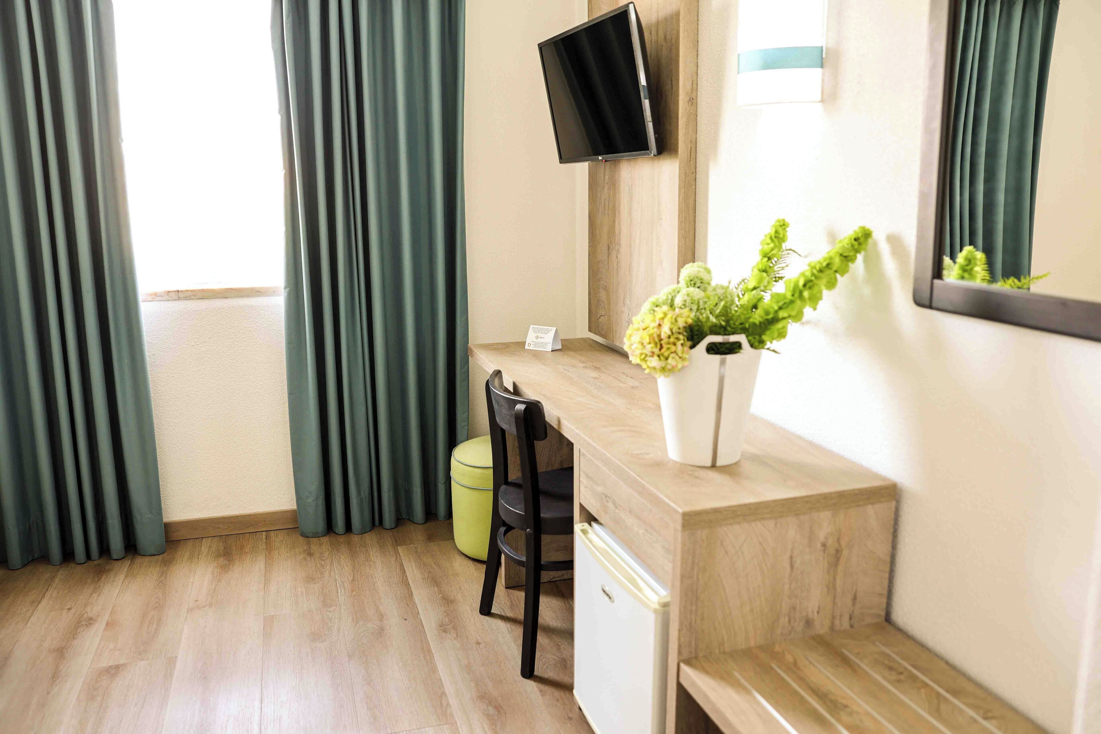 Habitación de hotel con piso de madera, un escritorio con silla, un refrigerador pequeño, un televisor montado en la pared y una planta en maceta sobre el escritorio.