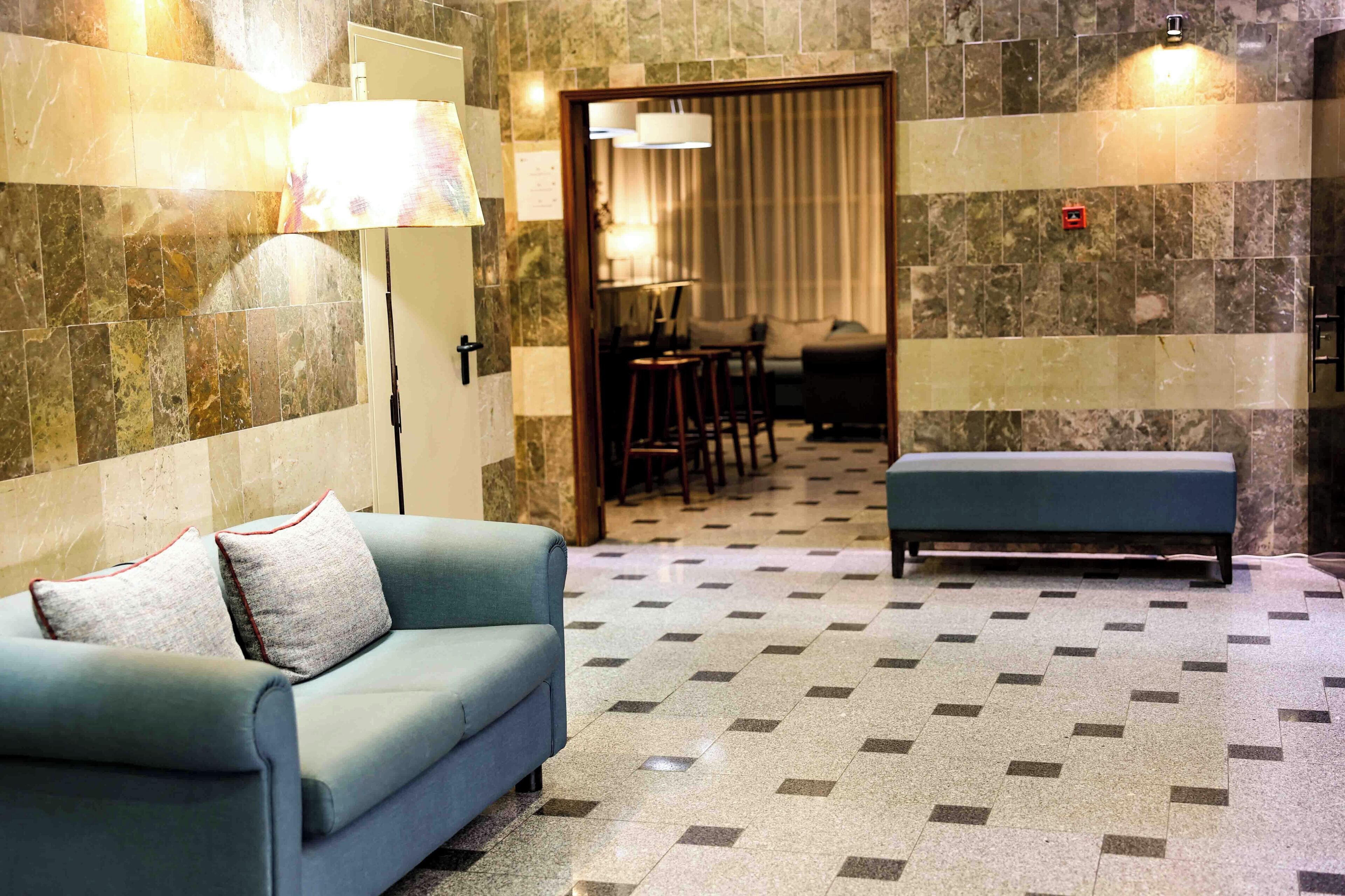 A hotel room with a sofa, a bench, a dining area with chairs and a table, and a patterned floor.