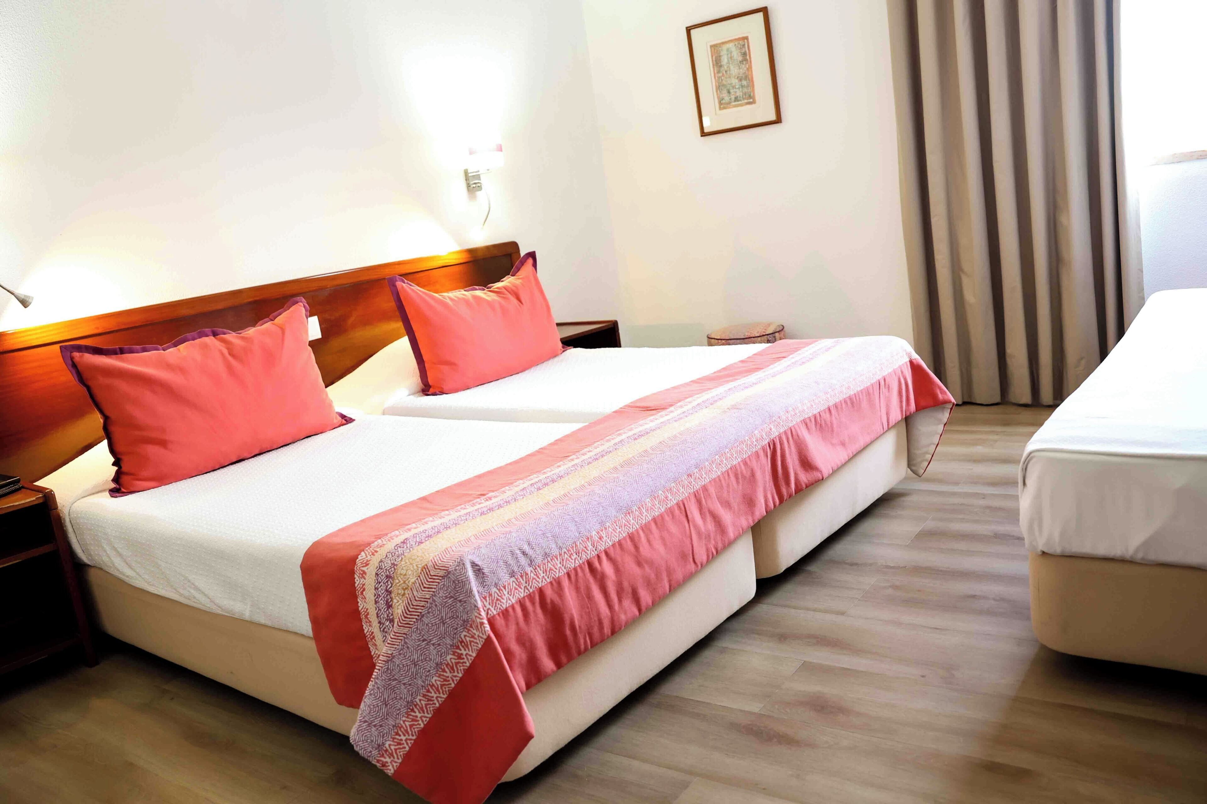 A hotel room with a double bed, a bedside table, a nightstand, a lamp, a window with curtains, and a framed picture on the wall.