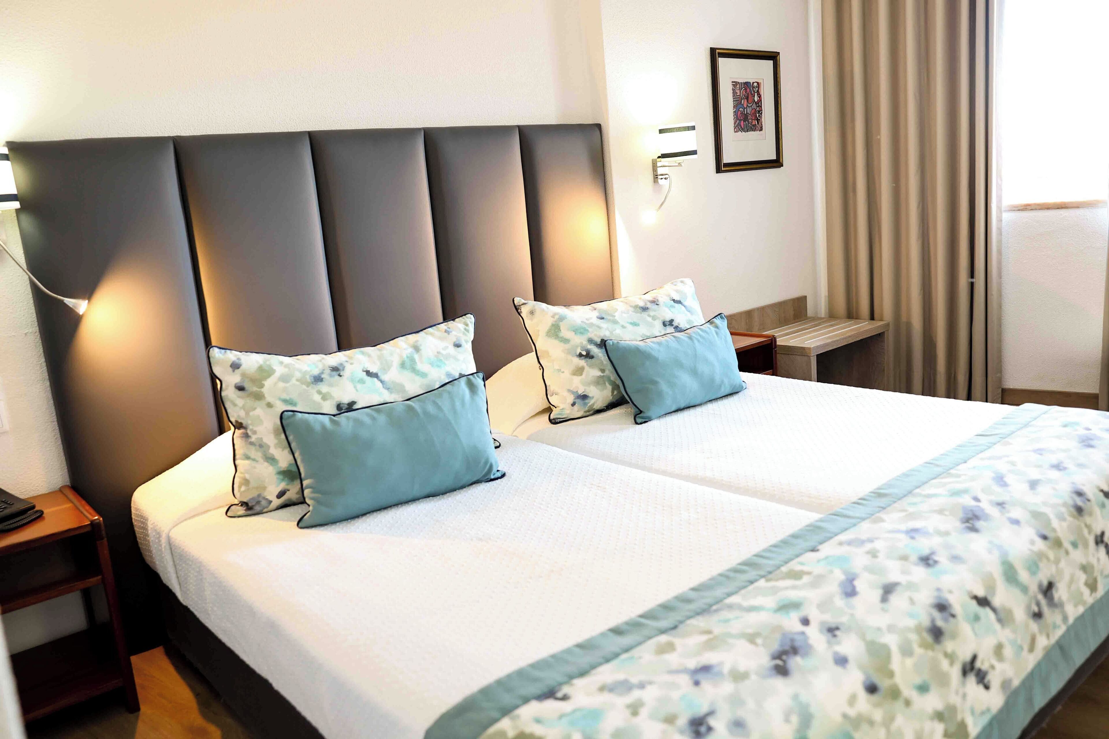 A double bed with two pillows, a bedside table, a nightstand, a wall-mounted lamp, and a framed picture.