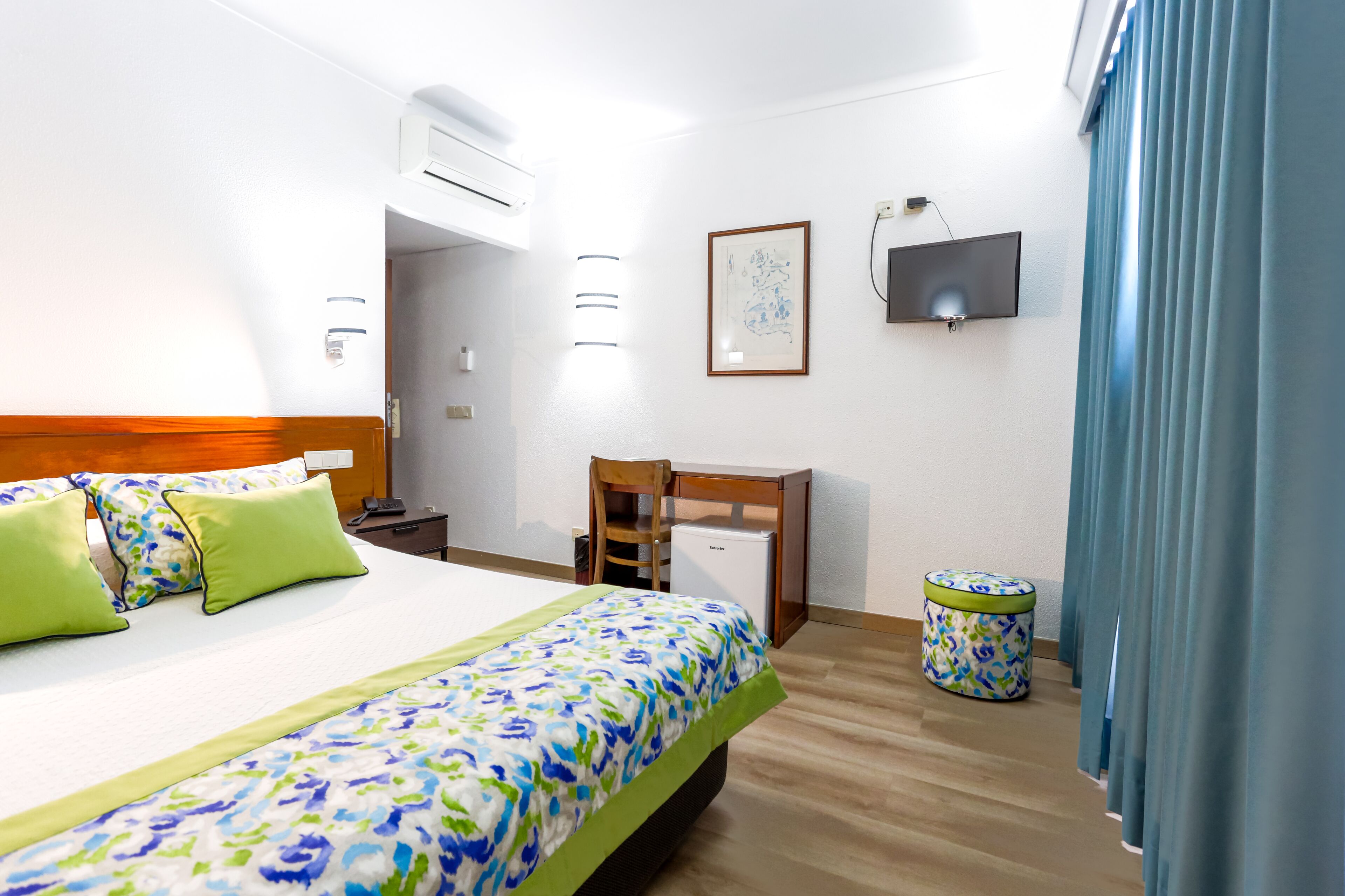 A hotel room with a bed, a desk, a chair, a TV, and a small table.