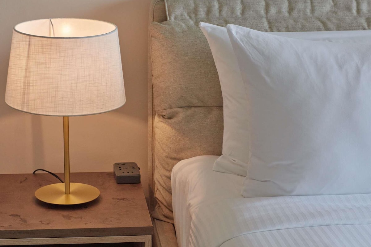 Un lit avec du linge de lit blanc, une lampe de chevet avec un pied doré et une multiprise sur une table de chevet en bois.