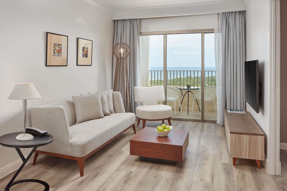 A modern living room with a sofa, coffee table, and chairs, a TV, and a balcony with a view.