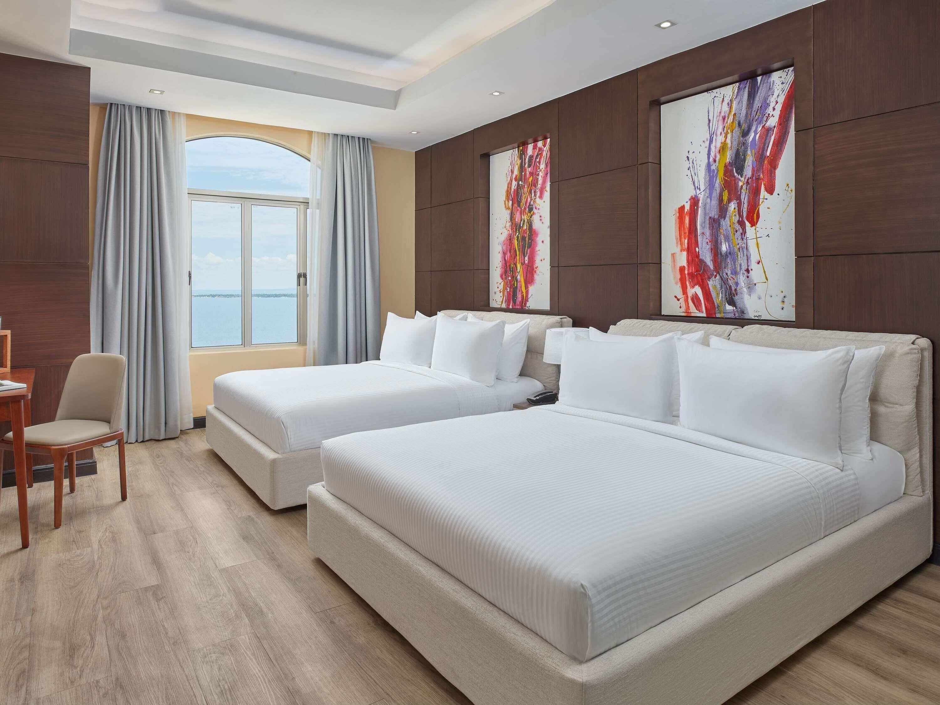 Habitación de hotel con dos camas, un escritorio y una silla. Cuenta con un ventanal con cortinas y cuadros en las paredes.