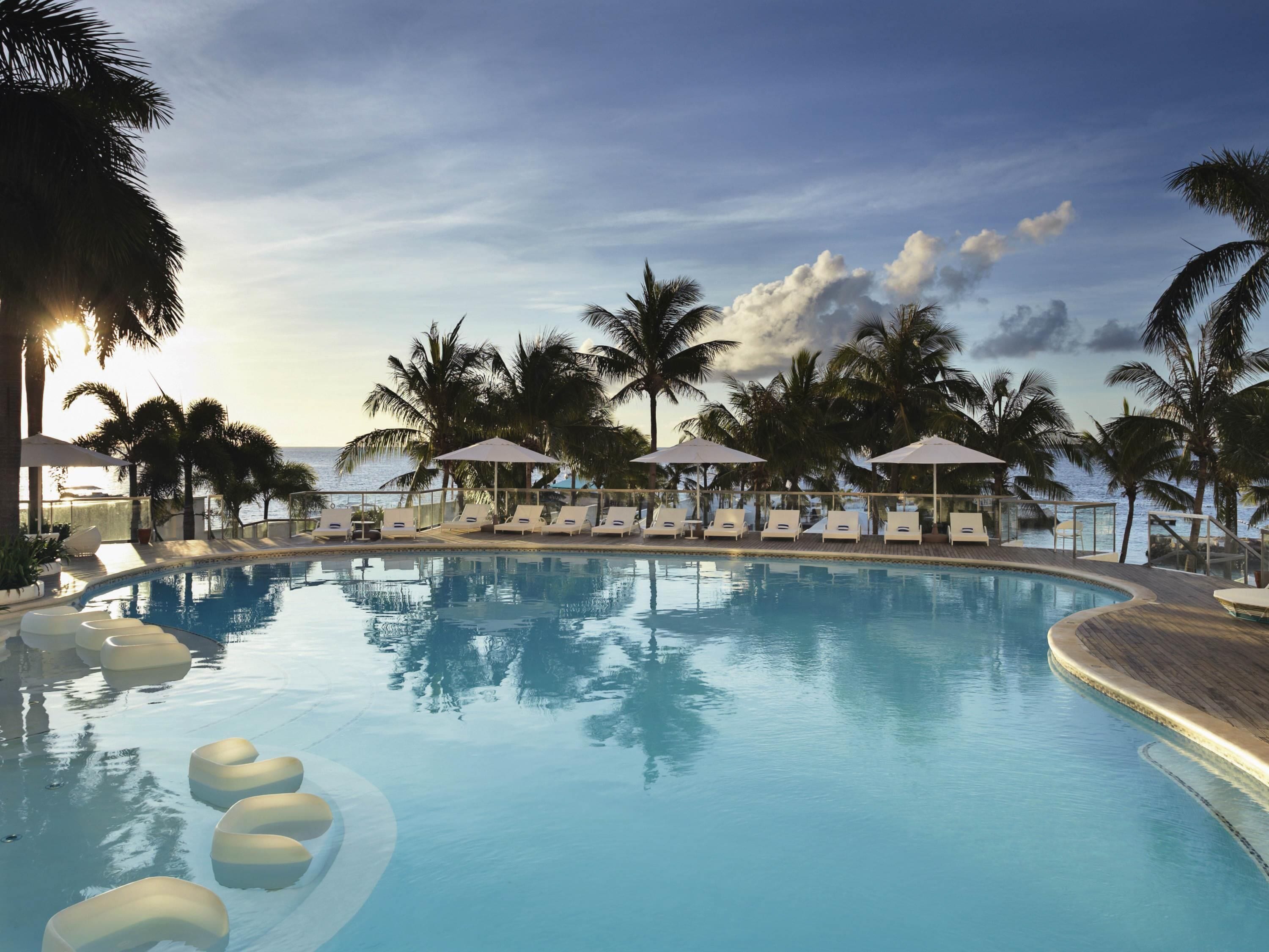 A pool with lounge chairs and umbrellas, surrounded by palm trees.