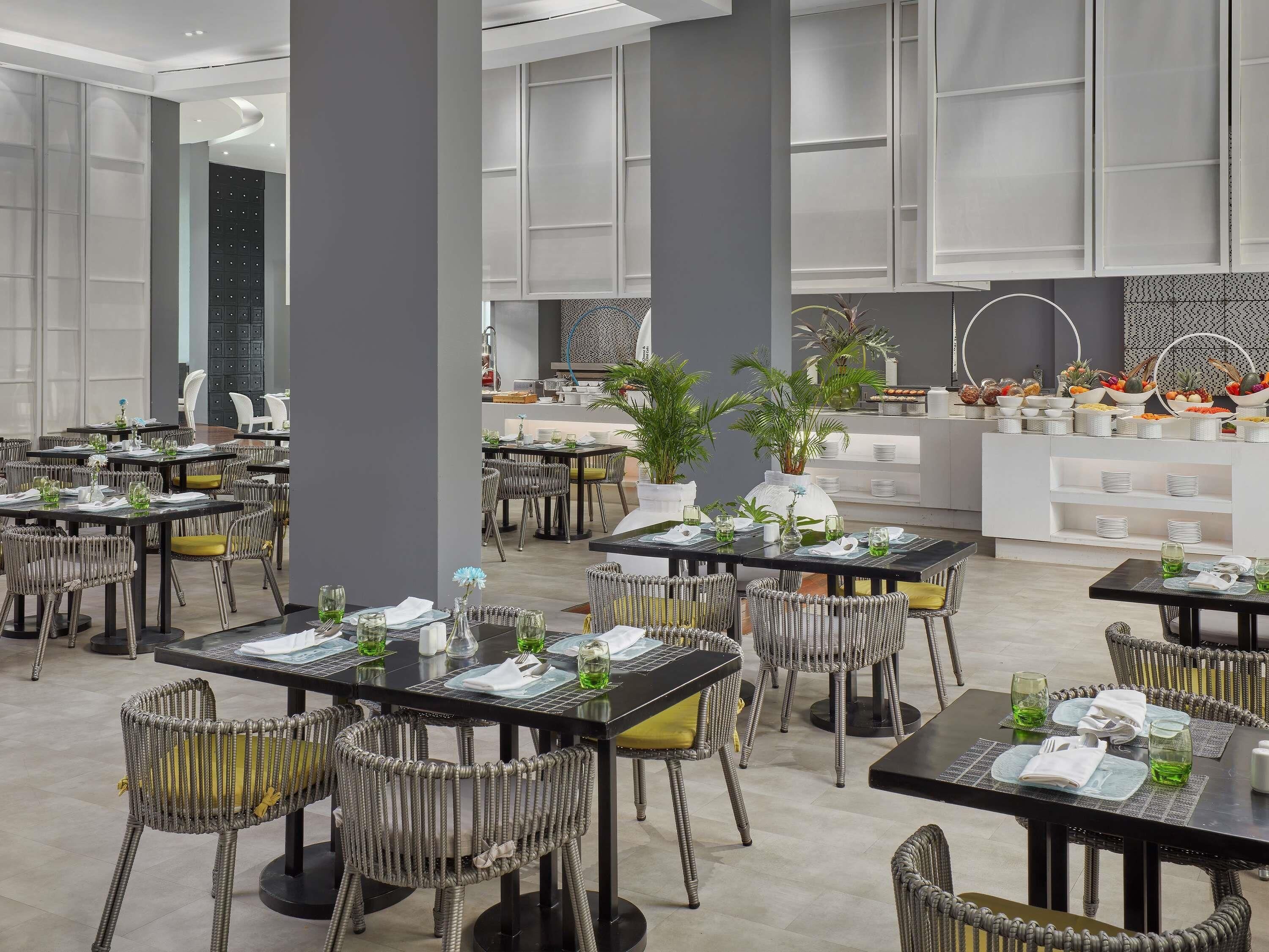 Een moderne eetruimte met meerdere tafels en rieten stoelen, met een buffet met diverse gerechten en decoratieve planten.