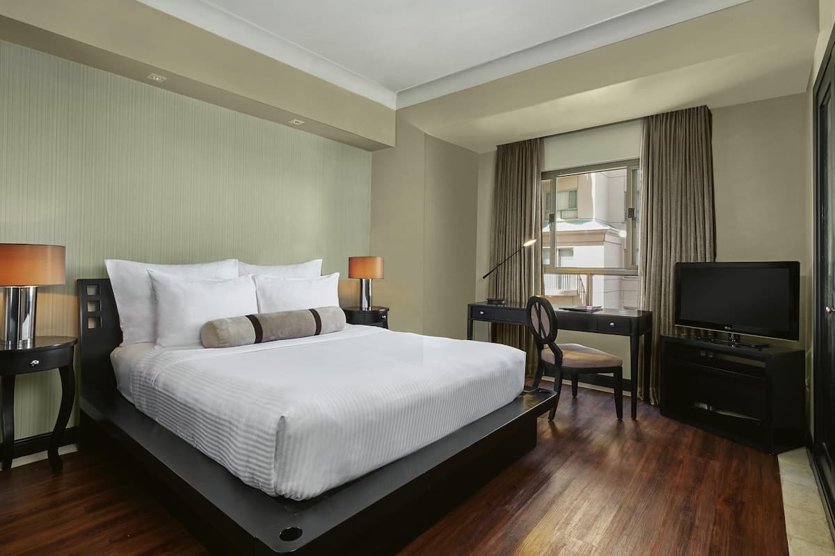 A hotel room with a large bed, two bedside tables with lamps, a desk with a chair, a television, and a window with curtains.