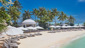 Une plage avec des chaises longues et des parasols, une structure en forme de dôme, des palmiers et un ciel bleu dégagé.