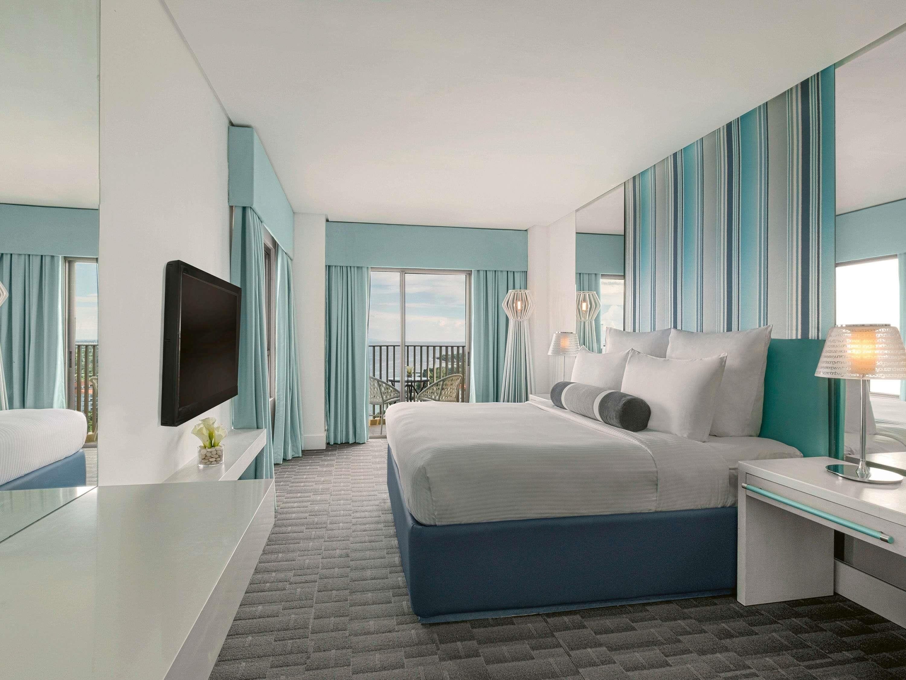 Habitación de hotel con cama, escritorio, televisión y balcón con vistas.