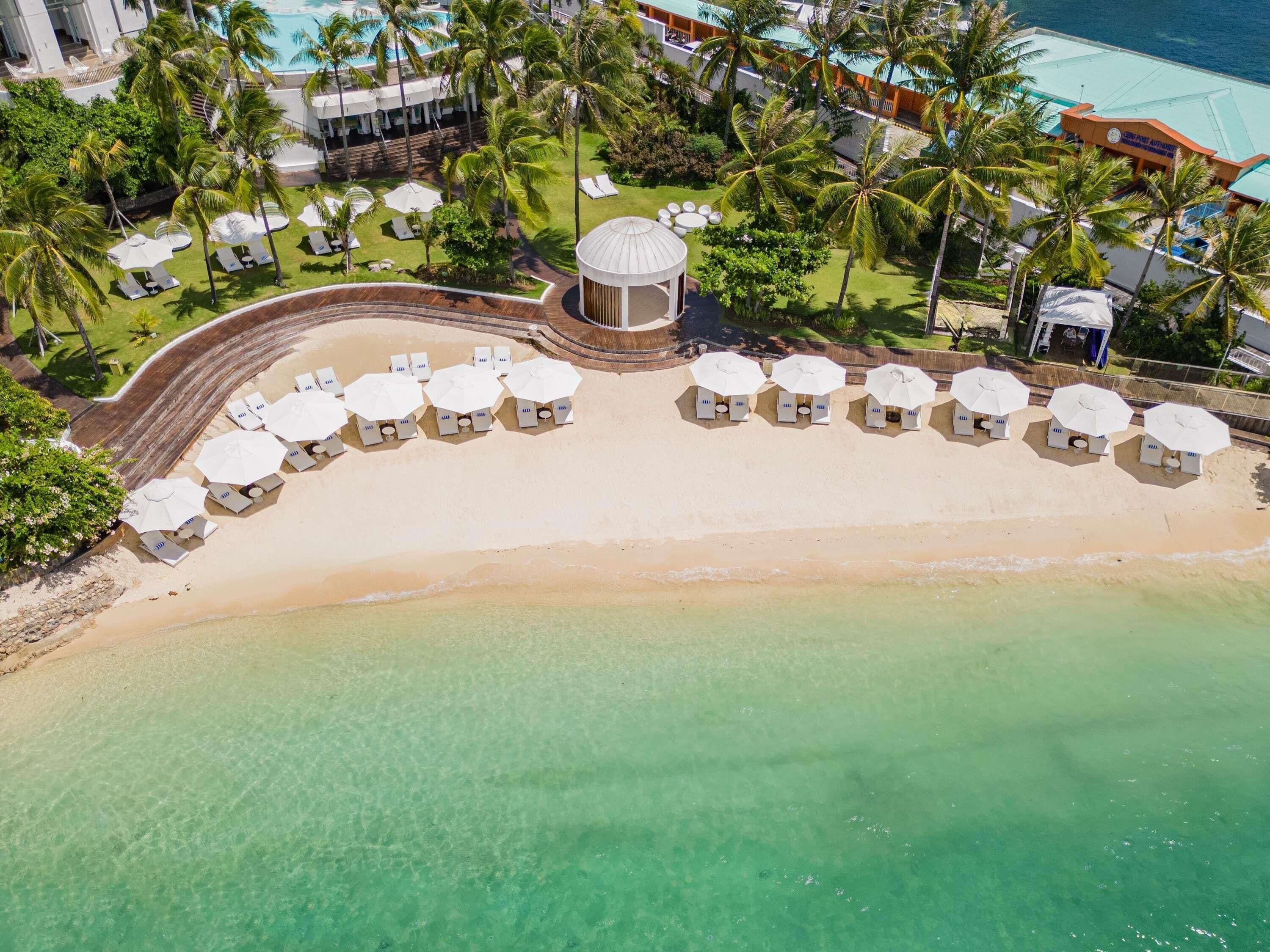 An aerial view of a beach resort with sun loungers and umbrellas, a pool, and palm trees.