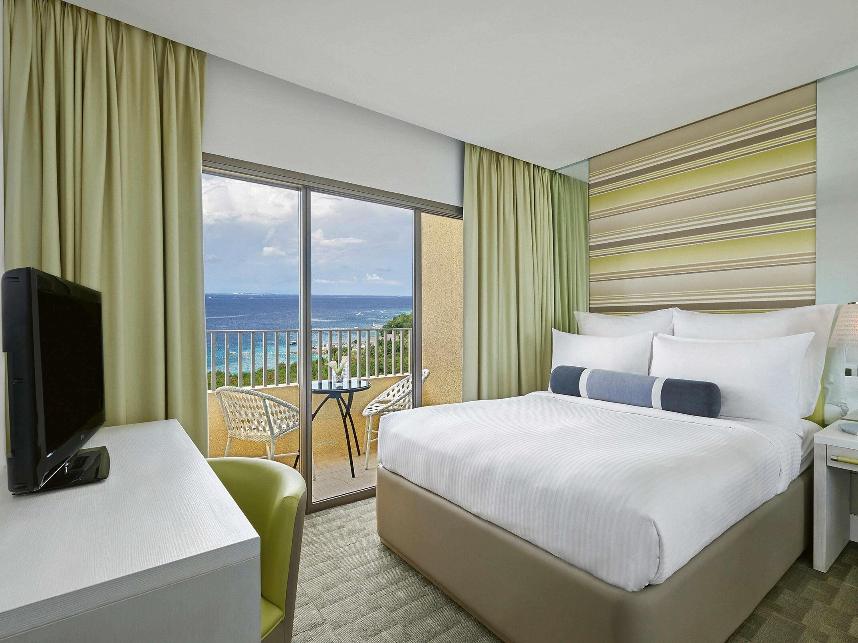 A hotel room with a bed, a desk, a TV, and a balcony with a view of the ocean.