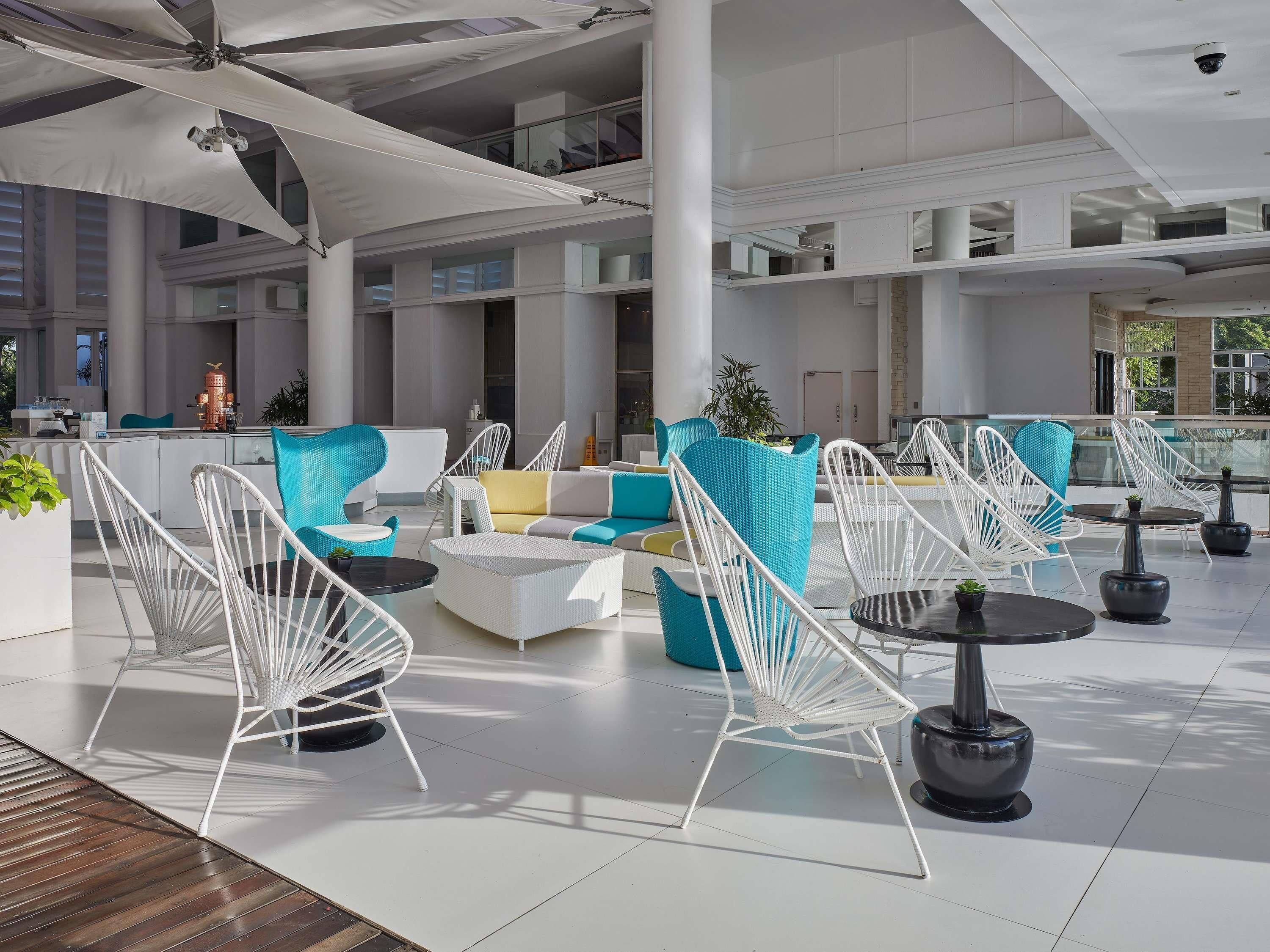 Un área de estar al aire libre moderna con mobiliario de mimbre blanco y azul, una estación de café y vistas del interior del edificio.