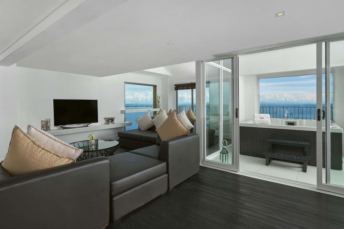 A modern living room with a large sofa, a flat-screen TV, and a balcony with a view of the sea.