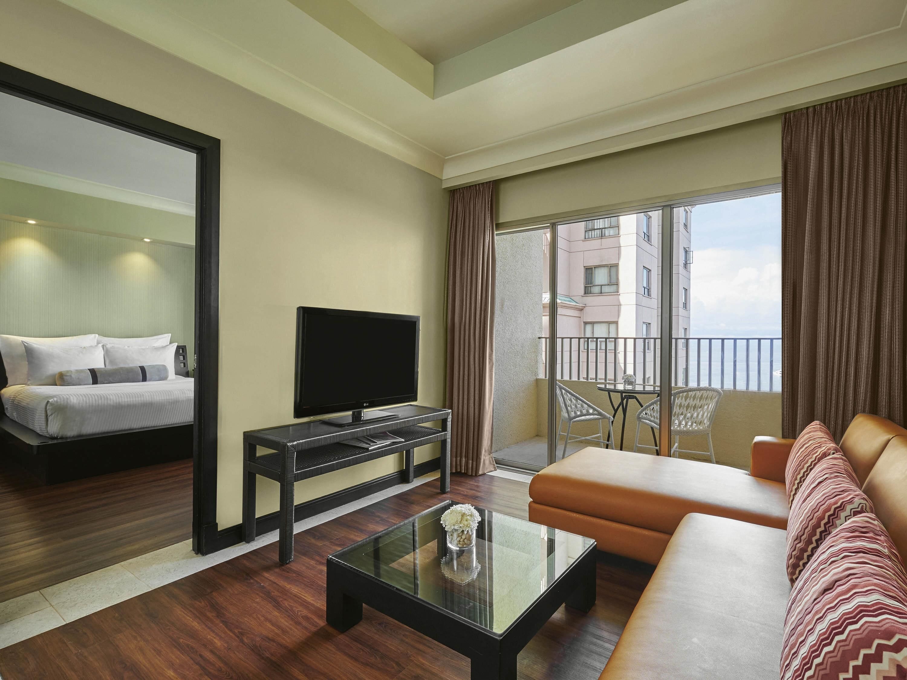 A hotel room with a bed, a television on a stand, a sofa, a coffee table, and a balcony with a view of buildings.