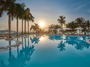 A pool with clear water reflecting palm trees and a sunset, surrounded by lounge chairs and umbrellas. - Mövenpick Hotel Mactan Island Cebu (Lapu-Lapu)
